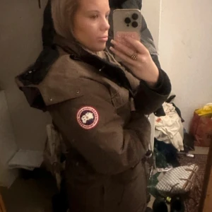 Brun parkajacka med huva - Säljer en brun parkajacka från Canada Goose med huva och emblem på ärmen. Jackan har en klassisk design med långa ärmar och flera praktiska fickor. Perfekt för kyliga dagar.