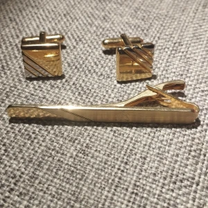 Guldfärgade manschettknappar och slipsnål - Elegant set med guldfärgade manschettknappar och en matchande slipsnål. Manschettknapparna har ett diagonalt randmönster och slipsnålen har en subtil design med en slät yta. Perfekt för att ge en stilfull touch till din outfit. Nytt skick, alldrig används. Ingen förpackning tyvärr.
