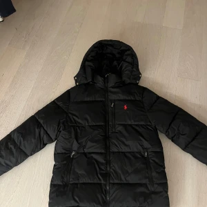 Svart dunjacka från Ralph Lauren - Säljer en svart dunjacka från Ralph Lauren med huva och dragkedja. Jackan har ett rött broderat märke på bröstet och praktiska fickor med dragkedjor. Perfekt för kyliga dagar. Finns inga defekter den är använd kanske 5 gånger. Hör av er vid frågor 🤝🏻