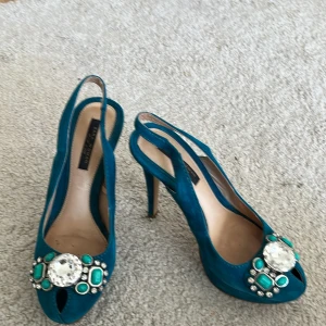 Turkosa klackskor från Zara Woman - Säljer ett par snygga turkosa klackskor från Zara Woman. De har en öppen tå och är dekorerade med stora glittriga stenar och turkosa pärlor framtill. Skorna är i mocka och har en elegant slingback-design. Perfekta för festliga tillfällen!