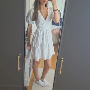 Vit omlottklänning från Gina Tricot - Säljer en superfin vit omlottklänning från Gina Tricot i storlek XS. Klänningen har korta puffärmar och en söt volangdetalj längst ner. Perfekt för sommarens alla fester eller en mysig dag på stan. Materialet känns lätt och luftigt, vilket gör den idealisk för varma dagar. 💖