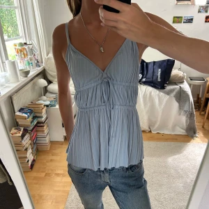 Ljusblå plisserad linnetopp - Säljer en superfin ljusblå linnetopp med smala axelband och v-ringning. Toppen har en plisserad design och knytning framtill som ger en söt detalj. Perfekt för sommardagar eller en avslappnad fest. Den är i bra skick och passar perfekt till jeans eller kjol.