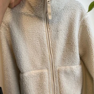 Beige teddyjacka med dragkedja - Mysig beige teddyjacka som är perfekt för kyliga dagar. Den har en dragkedja framtill och stora fickor för extra komfort. Jackan är supermjuk och ger en avslappnad look.