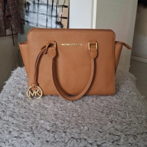 Brun handväska från Michael Kors - Säljer en snygg brun handväska från Michael Kors. Väskan har en elegant design med guldiga detaljer. Den har två handtag och en dragkedja upptill för säker förvaring. Perfekt för både vardag och fest! 👜✨obs axelbandet saknas, liten defekt enligt bild 
