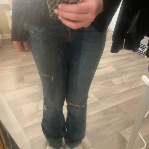 Blå jeans från Lindex - Säljer ett par blå jeans från Lindex i storlek 36. De har en cool, sliten look med revor på knäna och en bootcut-stil. Perfekta för en avslappnad vardagsstil. De är i välanvänt skick men fortfarande supertrendiga!
