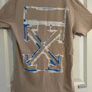 Säljer en beige t-shirt från Off White med ett stort grafiskt tryck i blått och vitt på baksidan. Framsidan har en enkel text i blått med plats och årtal. Perfekt för en avslappnad stil!