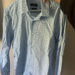 Skjorta Hugo boss - Säljer en snygg blå skjorta från Boss med ett diskret blommigt mönster. Skjortan är slim fit och har långa ärmar med knappar. 