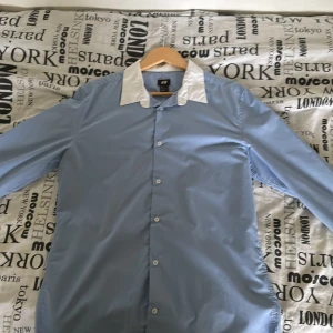 Blå skjorta från H&M - Säljer en stilren blå skjorta från H&M med vit krage. Skjortan har långa ärmar och knappar framtill. Perfekt för både vardag och fest!