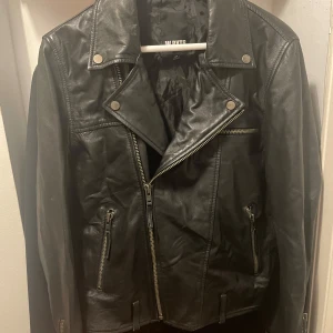 William Baxter Jacka Stevie Läder Jacka Svart - Skinnjacka i bikermodell från William Baxter. Jackan är tillverkad i ett mjukt och följsamt läder. Den har två dekorativa zippar vid ärmsluten och ett bälte i nederkanten. Två sidofickor samt en bröstficka med dragkedja. Nypris: 2500kr