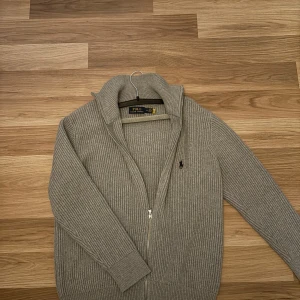 Grå ribbad tröja från Ralph Lauren - Säljer en stilren grå ribbad tröja från Ralph Lauren med dragkedja och hög krage. Tröjan har långa ärmar och en liten broderad logotyp på bröstet. Perfekt för kyligare dagar.