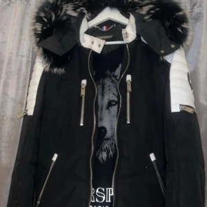 Svart och vit dunjacka med pälsdetaljer - Snygg svart dunjacka från Horspist med vita detaljer och en stor vargtryck på ryggen. Jackan har en avtagbar huva med svartvit päls och flera dragkedjor framtill. Perfekt för kalla dagar!