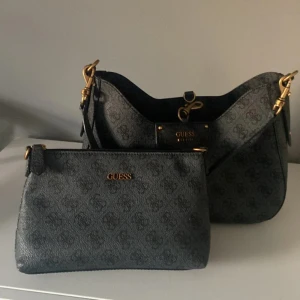 Grå blå axelväska från Guess - Snygg axelväska från Guess med guldfärgade detaljer. Väskan har en justerbar axelrem och en praktisk dragkedja. Inuti finns en liten väska som är som en clutch väska perfekt för fest. KÖPT FÖR 1600kr🩵