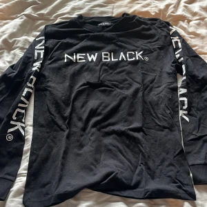 Svart långärmad tröja från New Black - Säljer en svart långärmad tröja från New Black med vit text på bröstet och ärmarna. Perfekt för en stilren och modern look. Tröjan är i storlek S och har en normal passform.