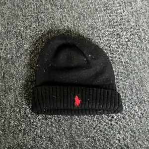 Svart mössa från Polo Ralph Lauren - Snygg svart mössa från Polo Ralph Lauren som inte är sliten bara lite smutsig men kan tvättas  med röd broderad logotyp framtill. Mössan är ribbad och tillverkad i ett mjukt material som passar perfekt för kyligare dagar. Den har en klassisk design som är lätt att matcha med olika outfits.