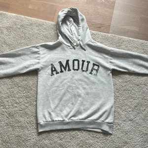 hoodie med amour tryck och stjärna - Säljer en grå hoodie från Gina Tricot med texten 'AMOUR' på framsidan och en stjärna på baksidan. Tröjan har en klassisk huva med dragsko och långa ärmar. Perfekt för en avslappnad stil.