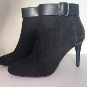 Svarta stövletter med klack och spänne - Eleganta svarta stövletter i mocka och skinn med spetsig tå och hög klack. De har en dekorativ rem med spänne runt ankeln och en praktisk dragkedja på sidan för enkel på- och avtagning. Endast använda en gång. Väldigt bekväma. Nypris 699 kr / 69€. Klackhöjd: 10cm