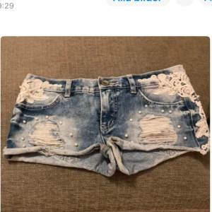 Blå jeansshorts med spetsdetaljer - Snygga blå jeansshorts med slitna detaljer och vacker vit spets på sidorna. Shortsen har pärlor som ger en extra touch av stil. Kort modell.