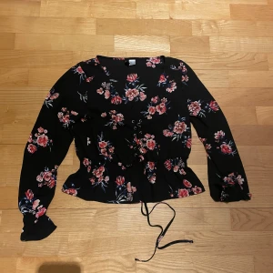 Blommig svart blus från Divided H&M - Säljer en svart blus från Divided med vackert blommönster i rött och rosa. Blusen har långa ärmar och en knytning i midjan för en fin passform. Perfekt för att ge en romantisk touch till din outfit.