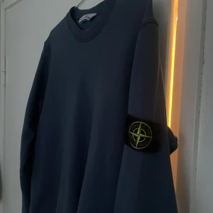 Blå tröja från Stone Island - Säljer en stilren blå tröja från Stone Island med deras ikoniska märke på ärmen. Tröjan har en klassisk rund hals och långa ärmar, perfekt för en avslappnad look. En liten defekt på framsidan men borde inte störa. Bra skick. S. Kvitto finns