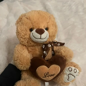 Brun nallebjörn med hjärta - Söt brun nallebjörn som håller ett hjärta med texten 'Love'. Den har en brun och vit rosett runt halsen och en broderad tass på ena foten. Perfekt som present eller dekoration.