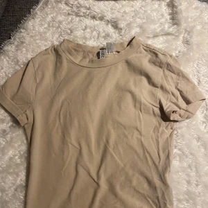 Beige t-shirt från H&M Divided - Säljer en enkel och stilren beige t-shirt från H&M Divided. Perfekt basplagg med korta ärmar och rund hals. Tillverkad i mjukt bomullsmaterial för en skön känsla. Passar till alla tillfällen och är lätt att matcha med andra plagg.