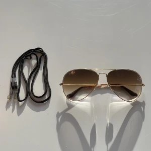 Ray-Ban pilotglasögon - Snygga aviator solglasögon från Ray-Ban med guldfärgad metallram och bruna tonade glas. Perfekta för en stilren look. Inkluderar ett svart glasögonband för extra säkerhet.