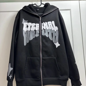 Svart hoodie med tryck - Säljer en svart hoodie med ett stort tryck på framsidan som säger 'Eternal Youth'. Hoodien har dragkedja och en luva med tryckdetaljer. Perfekt för en avslappnad stil.
