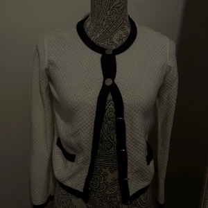 Vit kofta med svarta detaljer - Söt vit kofta med svarta kanter och knappar. Den har en rund halsringning och två fickor framtill.