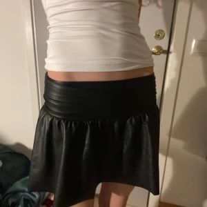 Svart kjol i skinnimitation - Snygg svart kjol i skinnimitation med en bred ribbad midja. Kjolen har en kort längd och en loose passform som ger en cool look. Perfekt för att ge din outfit en edgy touch.