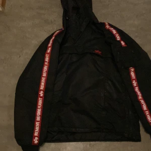 Svart jacka från Alpha Industries - Säljer en svart jacka från Alpha Industries med huva och coola röda detaljer längs ärmarna där det står 'REMOVE BEFORE FLIGHT'. Jackan har en avslappnad passform och är perfekt för en streetwear-look.