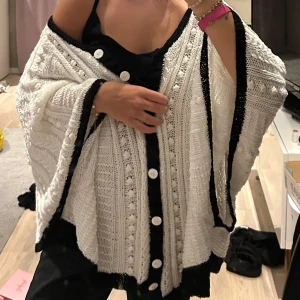 Vit stickad kofta med knappar - Säljer en mysig vit stickad kofta med svarta detaljer och knappar framtill. Perfekt för kyliga dagar och ger en stilren look. Koftan har en lös passform och är lätt att matcha med olika outfits.
