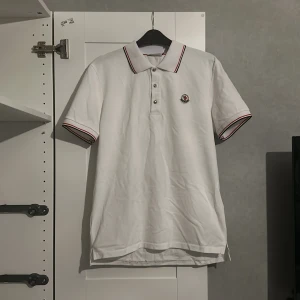 Vit pikétröja från Moncler - Säljer en stilren vit pikétröja från Moncler med klassisk krage och knappar framtill. Tröjan har röd och blå randdetalj på krage och ärmslut samt Moncler-logga på bröstet. Perfekt för en avslappnad men ändå elegant look.