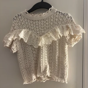 Vit spetsblus med volangdetaljer - Charmig vit spetsblus med vackra volangdetaljer över bröstet. Blusen har en fin stickad struktur och korta ärmar.
