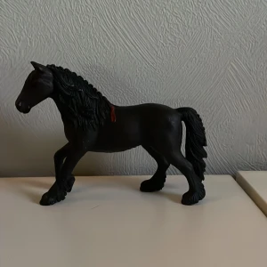 Schleich häst - En detaljerad svart hästfigur med flätad man och svans. Figuren är gjord av plast och har en realistisk design. Perfekt för samlare eller som dekoration.