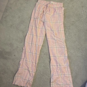Rosa rutiga byxor från Gina Tricot - Säljer ett par bekväma rosa byxor från Gina Tricot med rutigt mönster. Byxorna har en elastisk midja med snörning för justerbar passform. Perfekta för en avslappnad stil. Omsydda längst ner så de blir längre 