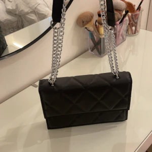 Svart quiltad axelväska med kedja - Snygg svart axelväska med quiltat mönster och en stilren kedja som axelrem. Perfekt för att ge din outfit en elegant touch. Väskan har en praktisk storlek för att bära dina nödvändigheter.