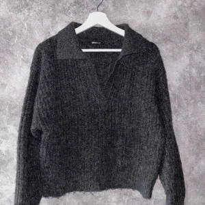 Mörkgrå stickad tröja från Gina Tricot - Mysig mörkgrå stickad tröja från Gina Tricot med en djup v-ringning och krage. Perfekt för kyliga dagar med sin mjuka och varma känsla. Tröjan har långa ärmar och en avslappnad passform.