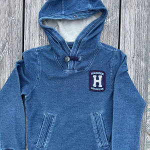 Blå hoodie från Hilfiger - Säljer en snygg blå hoodie från Hilfiger med en stor H-logga på bröstet. Tröjan har en knappdetalj vid halsen och snygga fickor som detalj framtill. Snygg denimblå nyans.  Perfekt för en avslappnad stil.