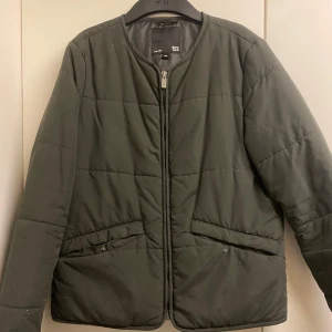 Grön quiltad jacka från H&M - Säljer en stilren grön quiltad jacka från H&M i storlek XS. Jackan har en dragkedja framtill och praktiska fickor med knappdetaljer. Perfekt för kyligare dagar med sin långa ärmar och bekväma passform.