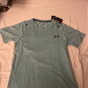 Under armour T-shirt  - Ljusgrön under armour T-shirt med reflekterande logga på bröstet och ett riktigt fett mönster över hela tröjan. Helt oanvänd. Tveka inte att höra av dig vid minsta fundering!