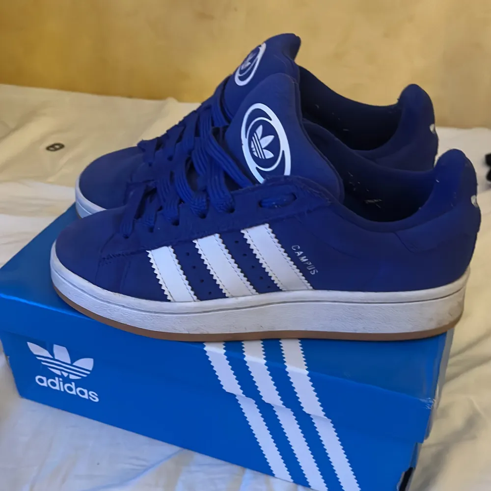Säljer ett par blå Adidas Campus sneakers med vita detaljer och klassiska tre ränder på sidan. Skorna har snörning och en bekväm Ortholite-innersula. Perfekta för en sportig och stilren look.. Kengät.