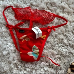 Röd stringtrosa med fjärilsmönster - Snygg röd stringtrosa med blommigt och fjärilsmönster. Den har knytband på sidorna och en vacker spetsdetalj upptill. Självklart oanvända 