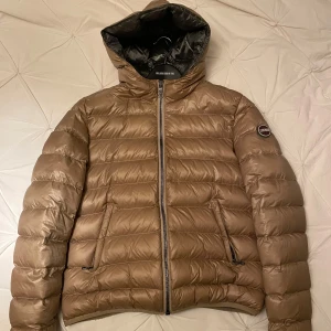 Brun/guld dunjacka från Colmar - Snygg brun/guld dunjacka från Colmar med dragkedja och huva. Jackan har en quiltad design och två praktiska fickor med dragkedja. Sparsamt använd. Som ny. Stlk40, som S/M