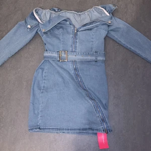 Jeansklänning med bälte - Snygg jeansklänning i blått med offshoulder-design och dragkedja framtill. Klänningen har långa ärmar och ett matchande bälte i midjan för en stilren look. Perfekt för en trendig outfit!
