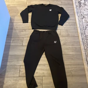Svart set SikSilk - Säljer ett stilren svart set med sweatshirt och byxor från SikSilk med deras logga på bröstet och benet. Tröjan har långa ärmar och ribbade muddar vid ärmslut och nederkant. Perfekt för en avslappnad stil. 🖤