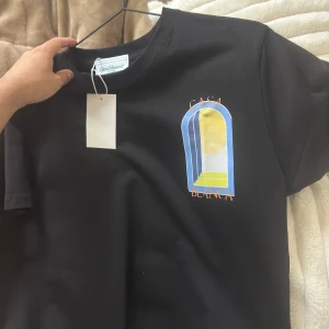  Casablanca t shirt - Snygg svart t-shirt från Casablanca med ett färgglatt tryck på bröstet som föreställer en stiliserad dörröppning i blått, gult och vitt. Perfekt för en avslappnad stil. Helt ny storlek Small kommer med tag säljs för 900kr 