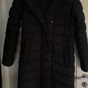 Svart lång dunjacka - Säljer en svart lång dunjacka med dragkedja och knappar. Jackan har en quiltad design och långa ärmar, perfekt för kalla dagar. Den är stilren och funktionell med fickor på sidorna.Didrikson i storlek 34, den är för stora för mig, skulle säga att dem nog passar upp till en liten 38/M beroende på vart man drar. Har knappt använt dem så den är som nya.  Yttermaterial 100% polyester.  Längd 121 cm rakt ner över ryggen Bröst bredd 50 cm  Tvättas 40 grader skontvätt 