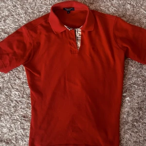 Burberry Piké [UTFÖRSÄLJNING av Plicksidan] - Snygg röd pikétröja från Burberry med klassisk krage och knappar. Detaljer i Burberrys ikoniska rutmönster vid knäppningen. Perfekt för en stilren look. Köpt på Vestiaire Collective därav real.