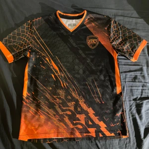 Svart och orange t-shirt med grafiskt mönster - Snygg svart t-shirt med orange detaljer och ett coolt grafiskt mönster. T-shirten har en v-ringad hals och korta ärmar med ett unikt tryck som ger en sportig look. Perfekt för dig som vill sticka ut!