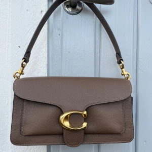 Brun axelväska i skinn med gulddetaljer - Coach tabby väska i skinn i färgen dark stone 🤎Knappt använd och därav mycket bra skick. Många fack för bra förvaring. Dustbag och långt axelband medföljer.